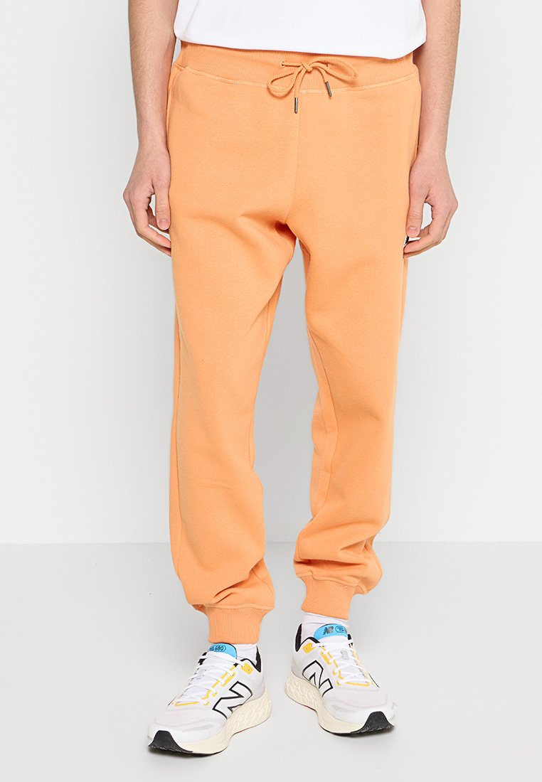 Pantalones de chándal de algodón naranja con cintura elástica y cordón, que cuentan con bolsillos laterales y puños acanalados. Llevados con zapatillas deportivas blancas.