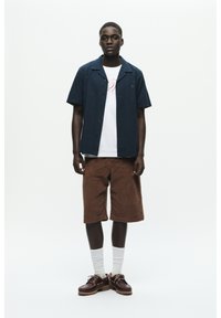 Cămașă bleu închis cu mânecă scurtă, tricou alb cu grafică dedesubt, pantaloni scurți din cordură maro, șosete albe și pantofi maro cu o talpă texturată.
