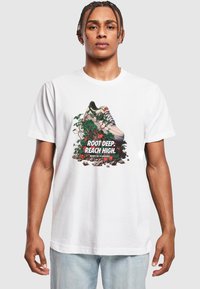 Bílá tričko s grafikou tenisky obklopené zelenými vinnými révami a kameny, s výrazným textem "ROOT DEEP. REACH HIGH."