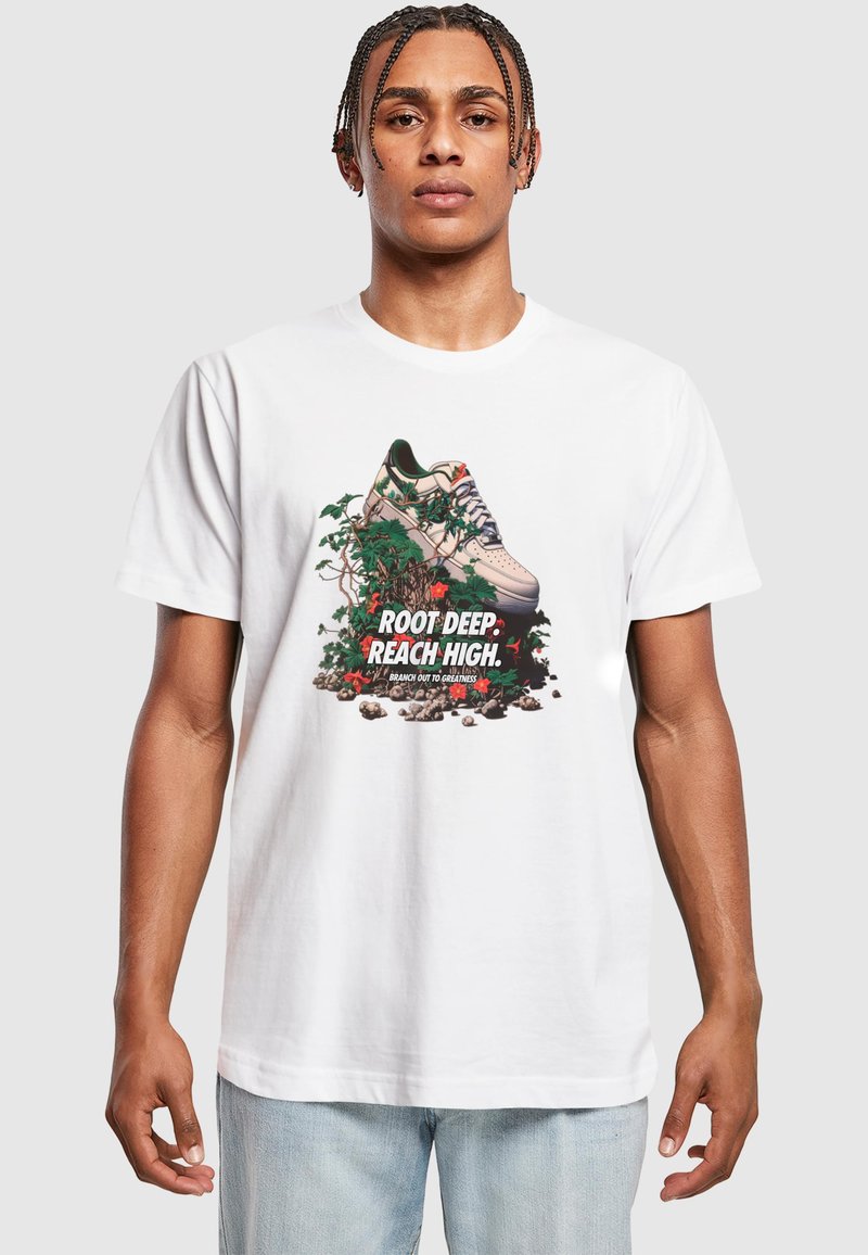 Bílá tričko s grafikou tenisky obklopené zelenými vinnými révami a kameny, s výrazným textem "ROOT DEEP. REACH HIGH."