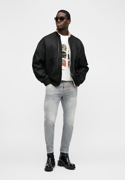 Homme portant une veste bomber noire, un t-shirt blanc avec un motif graphique, un jean gris clair usé, des bottes en cuir noires et des lunettes de soleil foncées, debout les mains dans les poches.