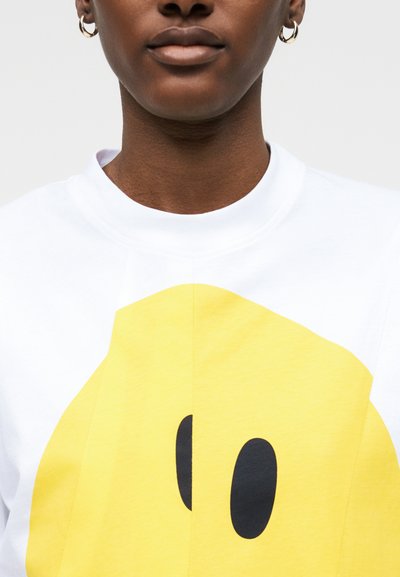 Άτομο που φοράει λευκό πουκάμισο με μεγάλο κίτρινο σχέδιο χαμογελαστού προσώπου και χρυσούς κρίκους στα αυτιά, εμφανιζόμενο από τον λαιμό μέχρι το στήθος.