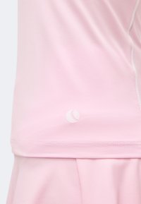 Ανοιχτό ροζ υφασμάτινη υφή με καμπυλωτή ραφή και μικρό λευκό κυκλικό λογότυπο κοντά στο τελείωμα, φορεμένο πάνω από ταιριαστά φαρδιά παντελόνια ή φούστα.