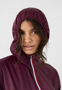 Giovane donna con lentiggini indossa una giacca con cappuccio color bordeaux chiusa con una zip, ciocche di capelli scuri sciolti incorniciano il suo viso su uno sfondo chiaro uniforme.