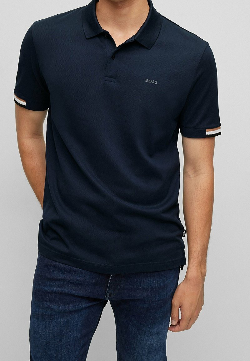 Homme portant un polo bleu marine foncé à manches courtes, avec des détails rayés sur les poignets et un petit logo "BOSS" sur la poitrine, associé à un jean foncé.