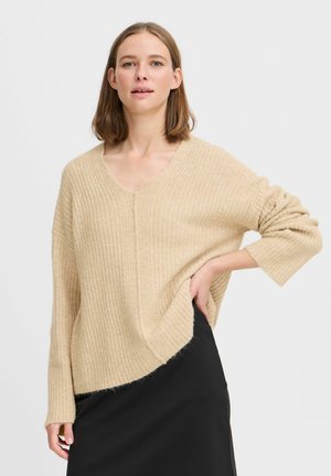 Pull beige côtelé avec un col en V et une coupe décontractée. Le tissu semble doux et texturé, avec un ourlet légèrement asymétrique et des manches larges.