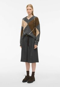 Pulover z V-izrezom in oversize slog z diamantnimi vzorci v sivi, rjavi in bež barvi; kombiniran s sivimi plisiranimi culottes hlačami in rjavimi čevlji na vezanje.