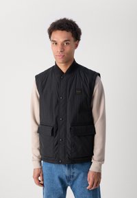 Schwarze ärmellose Weste aus strukturiertem Material, mit Druckknopfverschluss an der Vorderseite, zwei Taschen mit Klappen und einem kleinen Logo-Patch auf der Brust. Trägt man über einem beigen Langarmshirt.