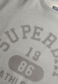 Sudadera gris de algodón, con un gran texto en gris oscuro. El texto incluye "SUPERDRY", "ATHLETE", "19" y "86" en un estilo negrita y colegial.