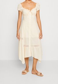 Woman wearing a cream, button-front midi dress with puff sleeves, lace details, and braided strap slip-on sandals against a plain background.