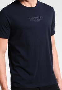 Muž v jednoduchém tmavě modrém tričku s krátkým rukávem, na kterém je na hrudi vytištěno "ARMANI EXCHANGE ICON" v světle šedé barvě.