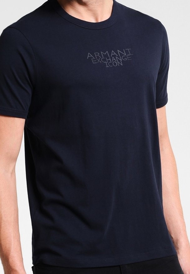 Muž v jednoduchém tmavě modrém tričku s krátkým rukávem, na kterém je na hrudi vytištěno "ARMANI EXCHANGE ICON" v světle šedé barvě.