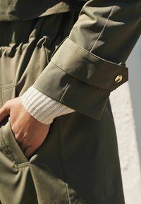 Oliwkowa zielona kurtka z szerokim mankietem ozdobionym złotym guzikiem, zestawiona z teksturowanym białym dzianinowym rękawem. Z przodu znajduje się kieszeń.