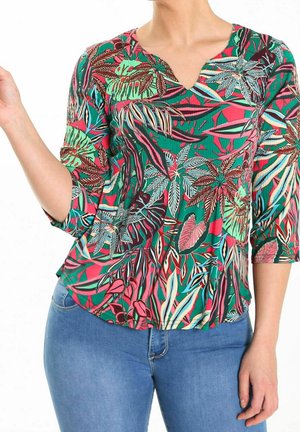 Kvinde iført en iøjnefaldende blomstermønstret top med trekvartlange ærmer kombineret med blå jeans, poserer mod en hvid baggrund.