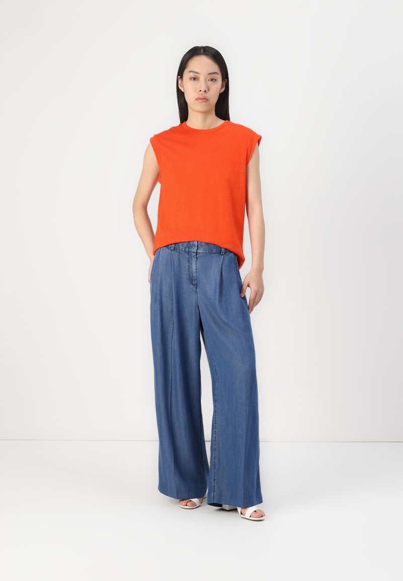 Haut rouge à manches courtes avec une coupe décontractée, associé à un pantalon en denim bleu à jambes larges. Design minimaliste, texture lisse et arrière-plan neutre.