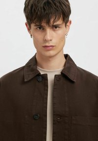 Brązowa bawełniana kurtka z kołnierzem, zapinana na guziki, z kieszenią na piersi, noszona na beżowej koszuli. Widoczna jest faktura tkaniny.