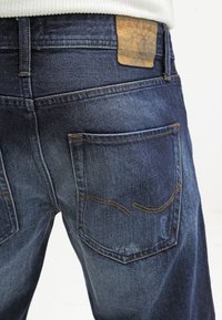 Dark blue denim jeans with faded areas, visible back pockets featuring curved stitching, and a worn leather brand label on the waistband.