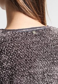 Tosig grå tröja med strukturerad stickning, försedda med en metallknapp i nackområdet och en kontrasterande halsringning.