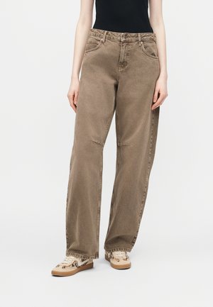 Άτομο που φοράει καφέ τζιν παντελόνι με φαρδιά μπατζάκια, μαύρο αμάνικο κορυφαίο και μπεζ αθλητικά παπούτσια με σκούρα σχέδια, στέκεται μπροστά σε λευκό φόντο.