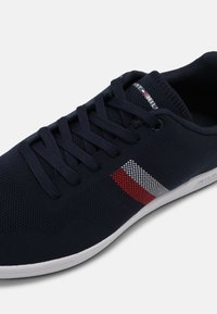Baskets bleu marine avec semelle blanche, texture en maille, lacets, et design rayé rouge-blanc-bleu sur le côté, logo Tommy Hilfiger sur la languette.