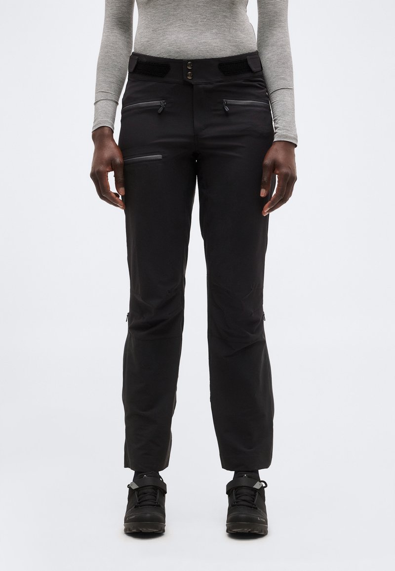 Osoba oblečená v čiernych nohaviciach so zipsom, sivom dlhom rukáve a čiernych turistických topánkach, stojaca pred svetlosivým jednoliatym pozadím.