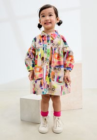 Vestito camicia colorato e decorato con colletto, maniche a sbuffo, tasche e bottoni; abbinato a scarpe da ginnastica bianche e calzini rosa.