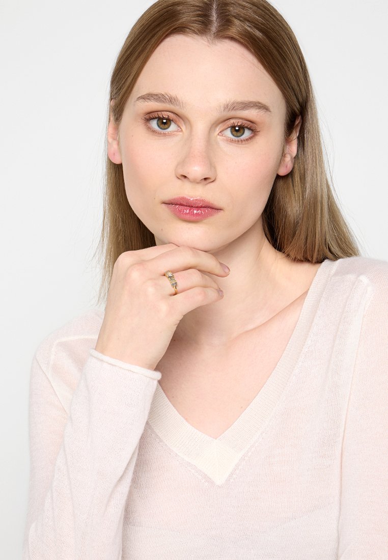 Jonge vrouw met lang lichtbruin haar die een lichtroze V-halstrui draagt, met een hand haar kin steunt en een gouden ring met heldere stenen toont.