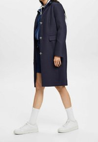 Cappotto lungo di un blu navy scuro con finitura texturizzata, dotato di bottoni argentati, tasche laterali e una vestibilità su misura, indossato sopra un completo blu e sneakers bianche.
