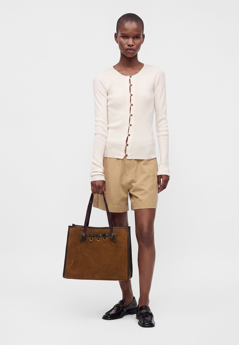 Femme portant un haut crème à manches longues boutonné, un short beige, des mocassins noirs, tenant un grand sac cabas marron avec des poignées foncées.