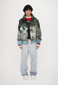 Fleece jacket with scenic print featuring greens and blues, black ribbed collar, and contrasting red and white striped cuffs. Worn over a black sweater and light wash baggy jeans.