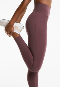 Egy személy lila, magas derekú leggingset visel, az egyik lábát hátranyújtja, míg a lábfejét fogja, fehér sportcipőben.