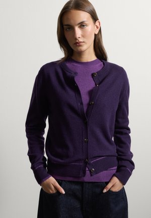 Junge Frau mit braunen Haaren, die eine dunkelviolette, geknöpfte Strickjacke über einem helleren violetten Pullover trägt, Hände in den Taschen dunkler Hosen, neutraler Hintergrund.