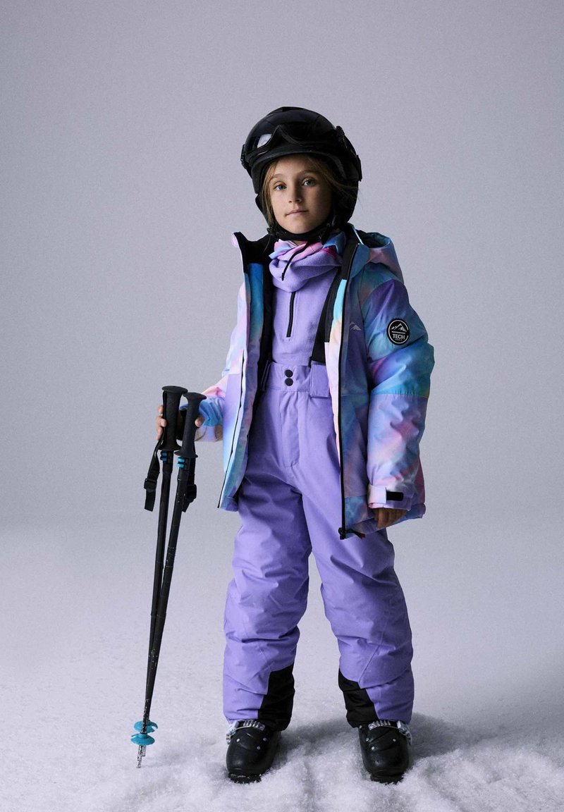 Krāsaina slēpošanas jaka ar gradienta rakstu, kopā ar lillā krāsas slēpošanas tērpu, melniem slēpošanas zābakiem un nūjām rokā. Aprīkots ar ķiveri.