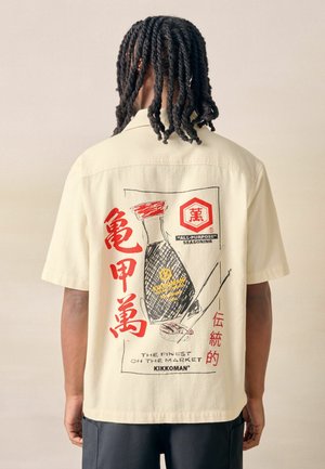 Punottu tukka ja beige paita, jossa kuvana Kikkoman-soijakastikepullo ja selässä punaisia japanilaisia merkkejä.