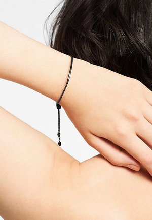 Zwarte leren armband met een strakke, gladde vormgeving, voorzien van een kettingdetail en een verstelbare sluiting, gedragen om een pols tegen de huid.