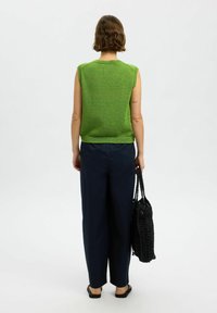 Grünes gestricktes ärmelloses Top mit Rundhalsausschnitt, kombiniert mit lockeren blauen Hosen. Schwarze gewebte Tasche wird in der linken Hand gehalten, Sandalen an den Füßen.