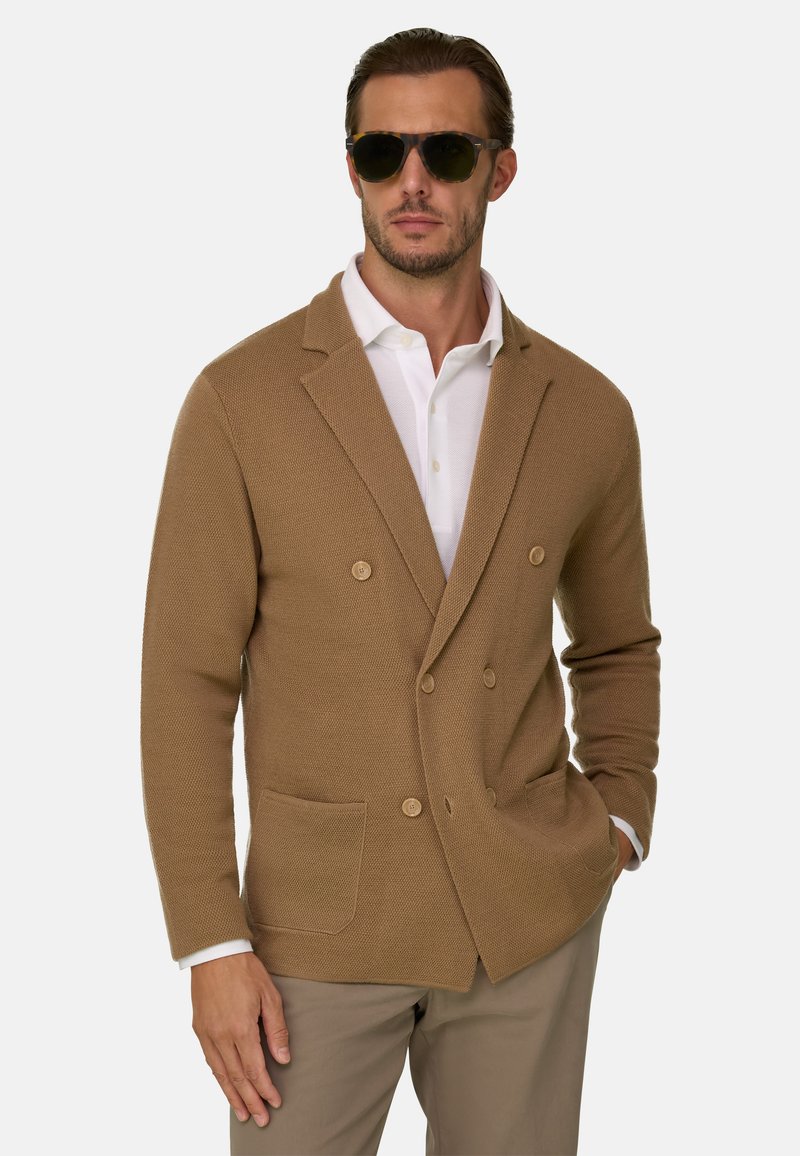 Homme portant des lunettes de soleil foncées, une chemise blanche à col, un blazer en maille marron à double boutonnage et un pantalon beige, debout avec une main dans la poche.