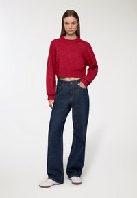 Donna che indossa un maglione rosso corto, jeans larghi scuri e sneakers bianche, in piedi contro uno sfondo bianco semplice.
