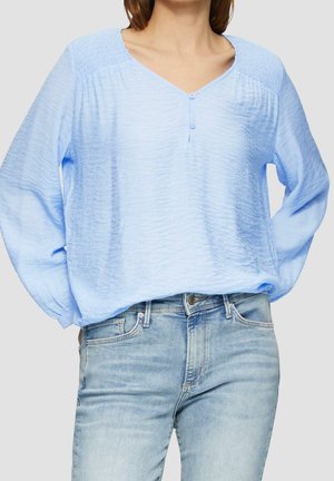 Kvinde iført lyseblå tekstureret bluse med knapper og lyseblå denimjeans, hænderne gemt bag ryggen.