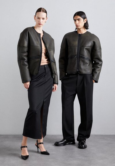 Zwei Models tragen olivgrüne Lederjacken mit Reißverschlüssen, kombiniert mit schwarzer Kleidung. Das Outfit der Frau umfasst einen Rock mit einem seitlichen Schlitz.