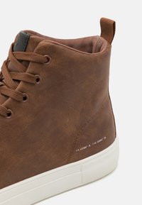 Brune faux skinnhøye sneakers med brune snørebånd, en teksturert krage og hvit gummisåle. Har trykte koordinater på siden.