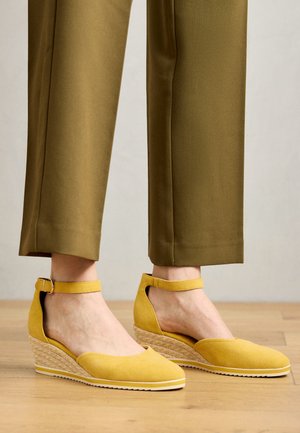 Sárga velúr magassarkú, bokapántos cipők, nyitott oldalúak, kötél mintázatú sarokkal, olajzöld nadrággal párosítva, fából készült padlón.