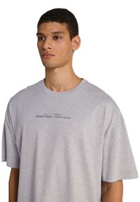 Ung mand med kort mørkt hår iført en lys grå oversized T-shirt med teksten "OFF DUTY GEAR HALO" på brystet.