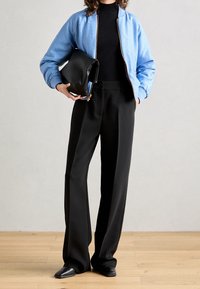 Chaqueta bomber azul claro con patrón texturizado, suéter negro de cuello alto, pantalones anchos negros y bolso de mano negro. Zapatos negros de punta.