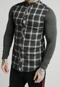 Camisa ajustada de manga larga con un patrón de cuadros en negro y blanco en la parte delantera y mangas grises lisas, con un diseño de botones y dobladillo redondeado.