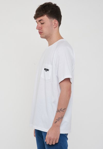 T-shirt in cotone bianco con taschino sul petto e logo nero. Vestibilità rilassata e maniche corte, con un tatuaggio a forma di foglia visibile sull'avambraccio.