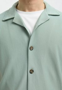 Maglietta a coste verde chiaro con colletto a punta, caratterizzata da due bottoni marroni e una camicia interna bianca. Il tessuto ha un aspetto granuloso.