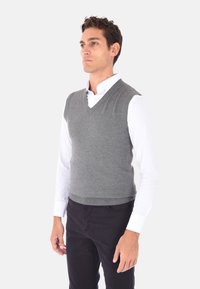 Gilet grigio lavorato a maglia con scollatura a V, indossato sopra una camicia bianca. Presenta una forma aderente e orlo a coste. Sono visibili pantaloni neri.
