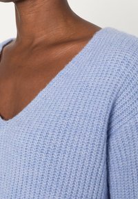 Light blue, knitted sweater with a V-neck design. Soft texture with visible stitching and slightly elongated sleeves.