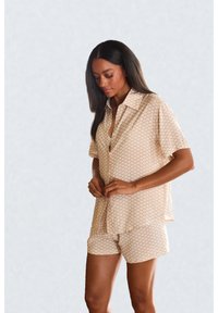 Žena s dlhými tmavými vlasmi zapína béžovú vzorovanú košeľu s krátkym rukávom a k nej vzorované krátke nohavice na jednoduchom svetlom pozadí.